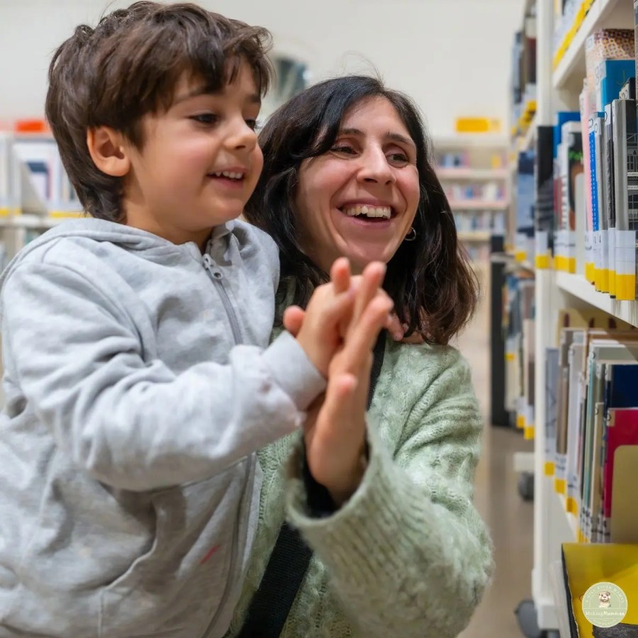 Mother and young child smiling together in a library aisle while looking at books on the shelves.
