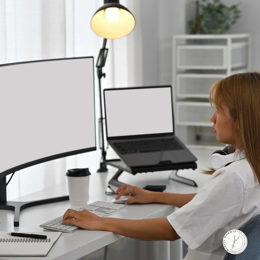 Woman working at a white desk with a curved external monitor and laptop setup, wearing headphones around her neck and a cup of coffee nearby, representing a modern home office workspace.