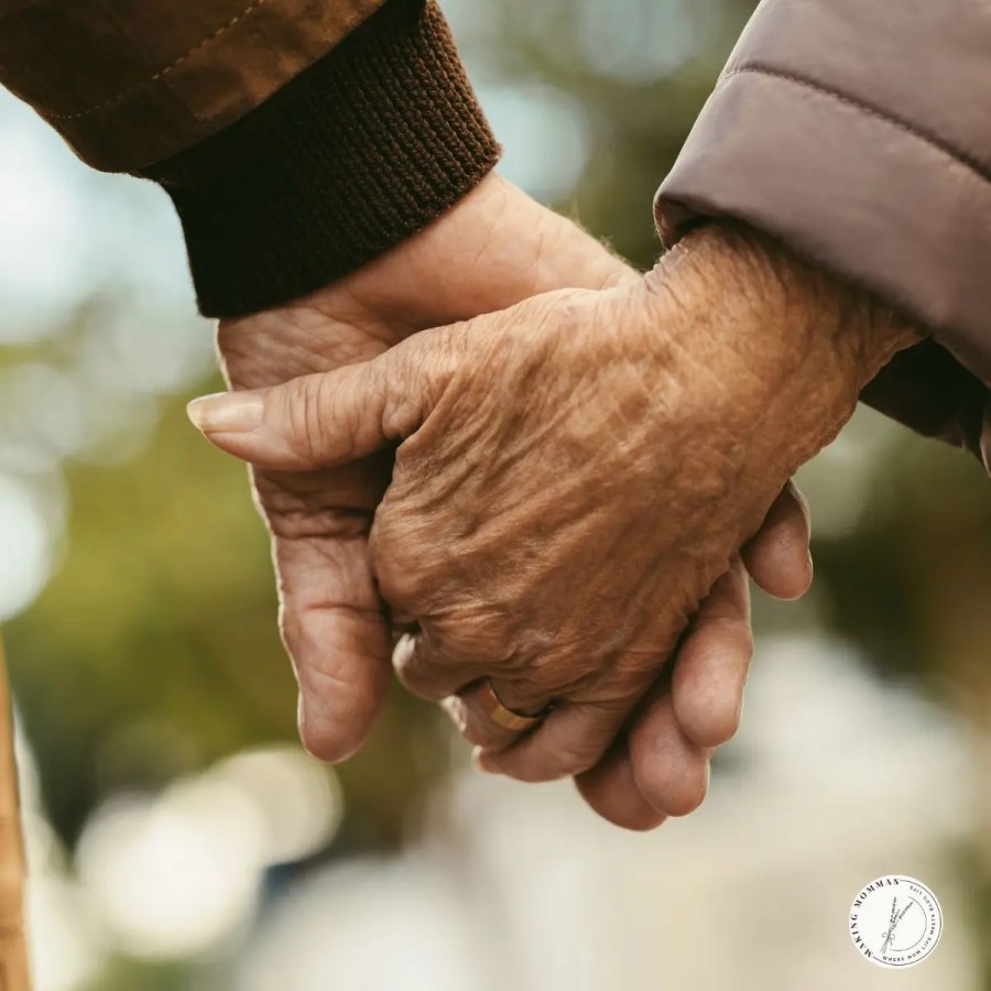 Close-up of an elderly couple holding hands, showing wedding rings and weathered hands, symbolizing lifelong love and commitment.