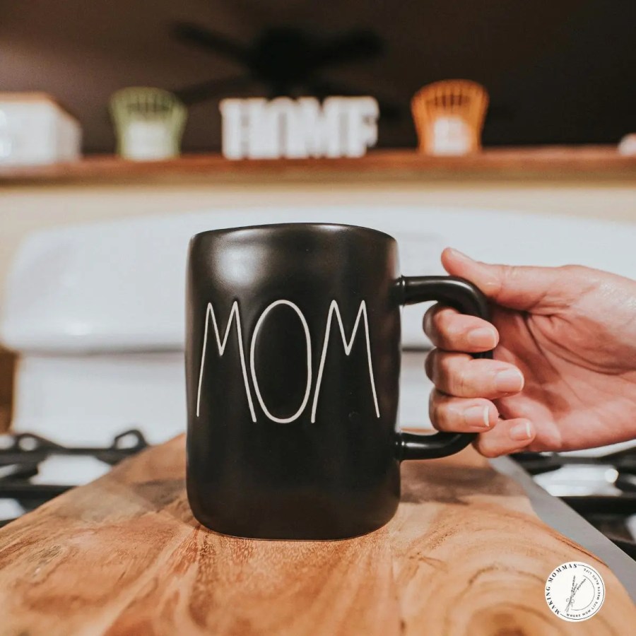 A hand holding a black ceramic mug with the word “MOM” written in white letters, resting on a wooden countertop in a cozy kitchen.