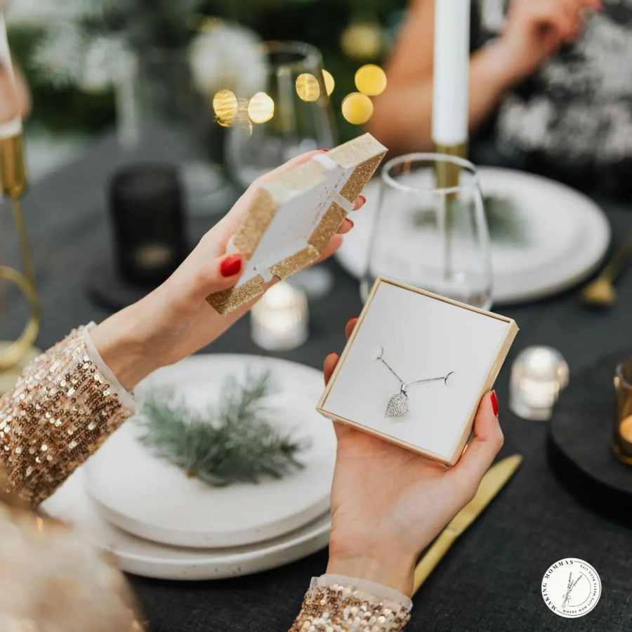 A woman opening a gold gift box at a festive dinner table, revealing a silver necklace with a pendant inside, surrounded by candles and holiday decor.