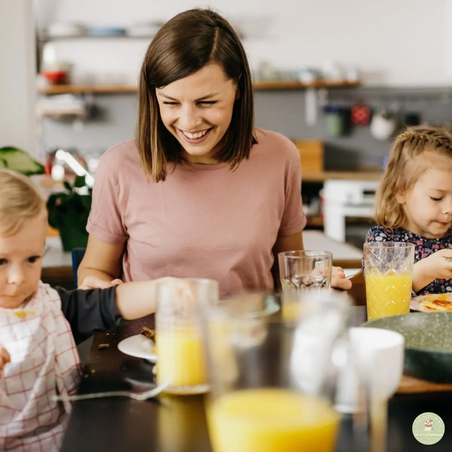 A mom with short brown hair, wearing a pink t-shirt, smiles happily while sitting at a dark dining table with two young children. To her left, a toddler boy with a white and red checkered bib is in the foreground, eating. To her right, a young girl with blonde pigtails and a floral dress is also eating. Glasses of orange juice and water are on the table, with a blurred kitchen in the background.