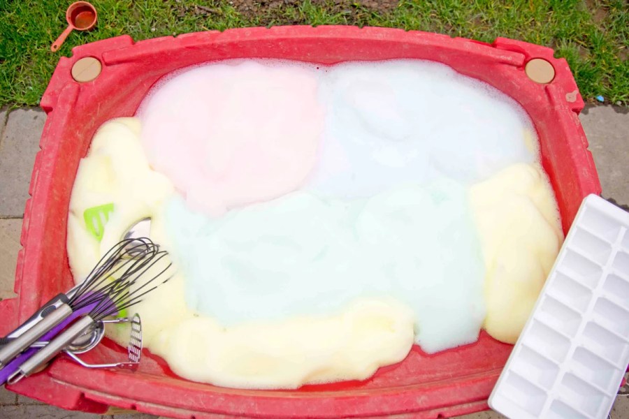 Outdoor sensory play setup with a plastic bin filled with colorful foam bubbles, along with kitchen tools like whisks and scoops for kids to explore and play.