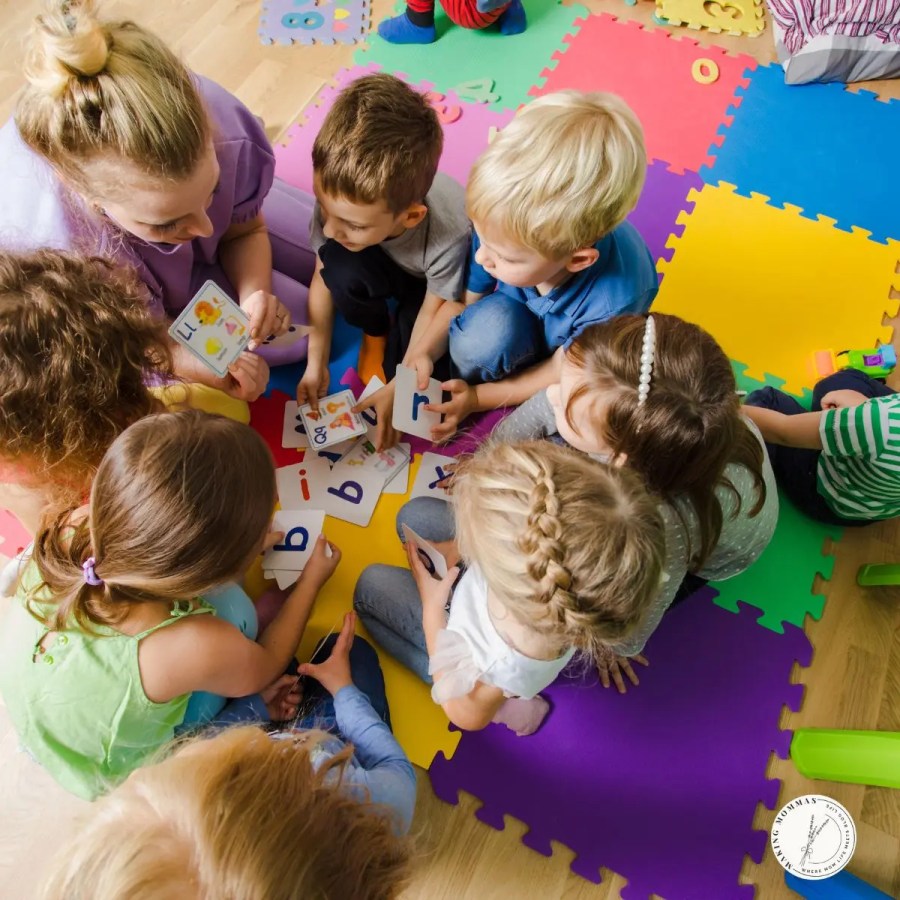 Preschool teacher sitting on colorful foam mats teaching a group of young children with alphabet flashcards during a structured learning activity.