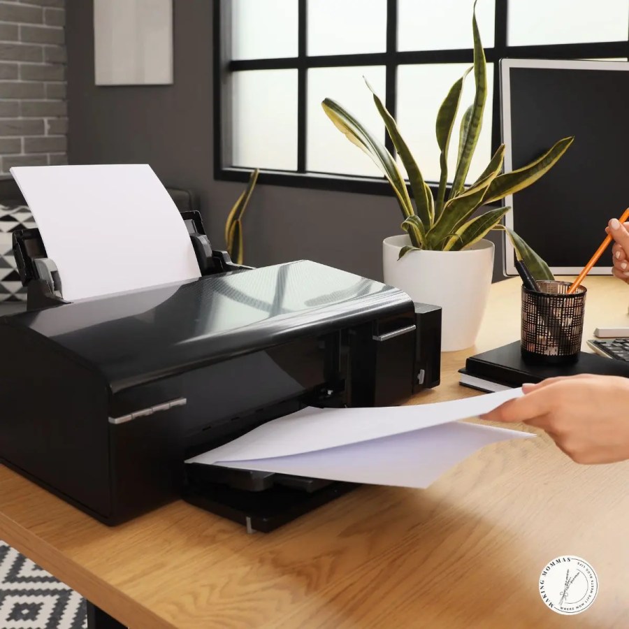 Close-up of a person loading paper into a black printer on a modern wooden desk with a potted plant, laptop, and pencil holder in the background, symbolizing a functional home office setup.