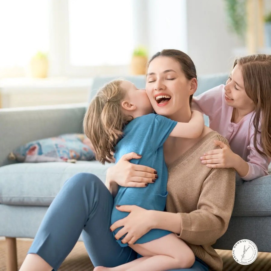 Happy mother sitting on the floor, hugging and laughing with her two young children in a cozy living room setting.