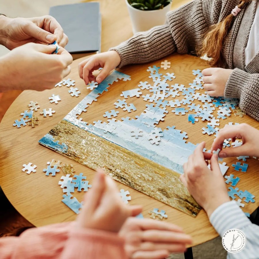 A family gathered around a wooden table working together on a scenic jigsaw puzzle with pieces of sky and sand.