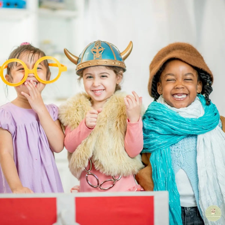 Three smiling young children dressed in playful costumes with oversized glasses, a Viking helmet, scarves, and dress-up accessories during pretend play.