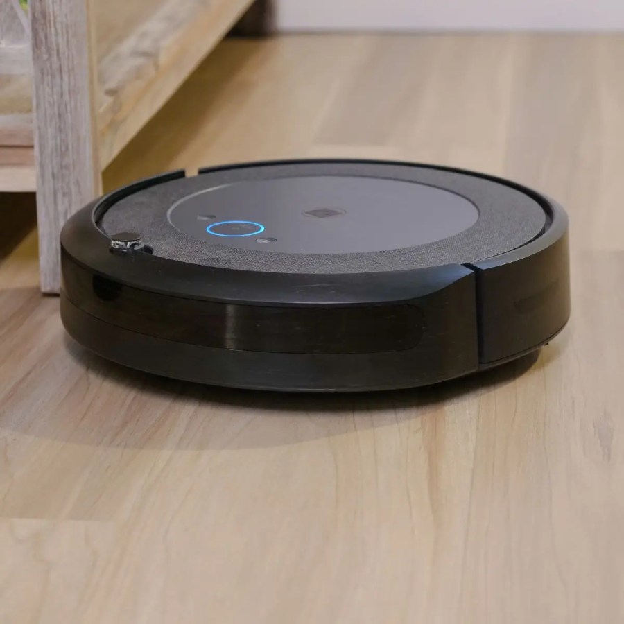 Alt text: A black robot vacuum cleaner moving across a hardwood floor next to a wooden cabinet.