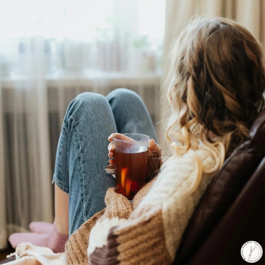 A mom sitting quietly by a window, holding a warm drink and taking a moment to rest and recharge during a calm part of the day.