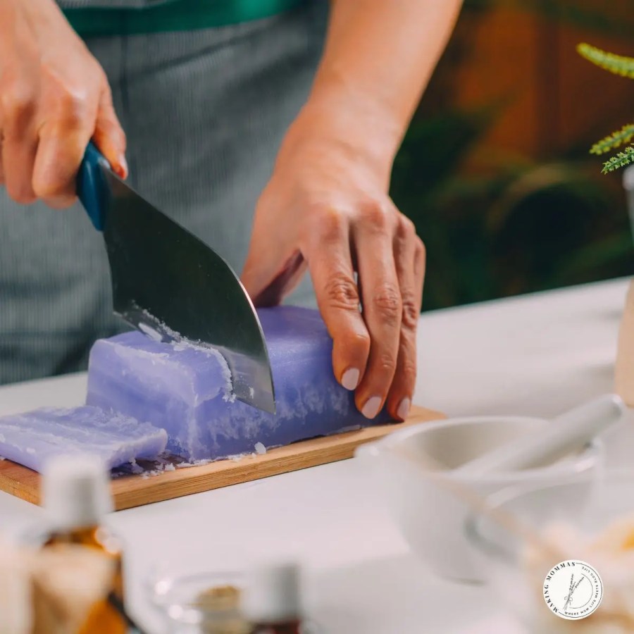 A person cutting a block of purple homemade soap on a wooden board with a soap cutter, surrounded by natural ingredients and bottles for soap making.