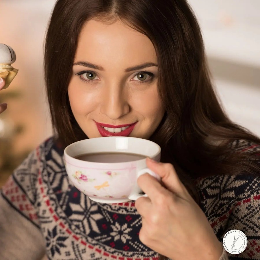 Woman in a festive winter sweater smiling while holding a floral teacup and enjoying a warm drink, capturing a cozy and cheerful holiday moment.