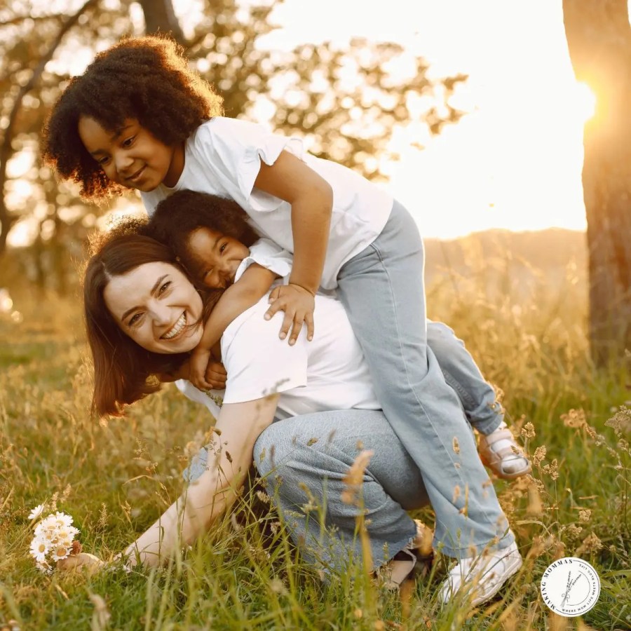 A smiling mom kneeling in a grassy field at sunset with her two young children climbing on her back, enjoying playful quality time together outdoors.