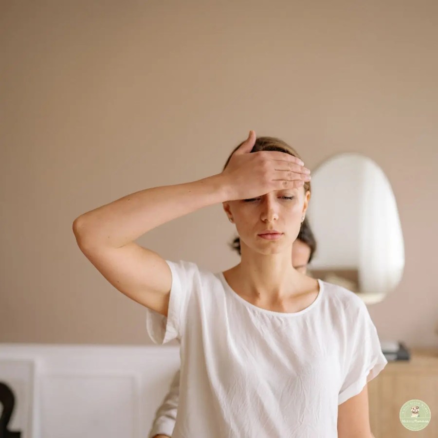 A young woman with light brown hair pulled back, wearing a simple white t-shirt, stands with her eyes closed and her right hand resting on her forehead. She appears to be feeling unwell or stressed. The background is a plain beige wall, with a partial view of an oval mirror and a person behind her.