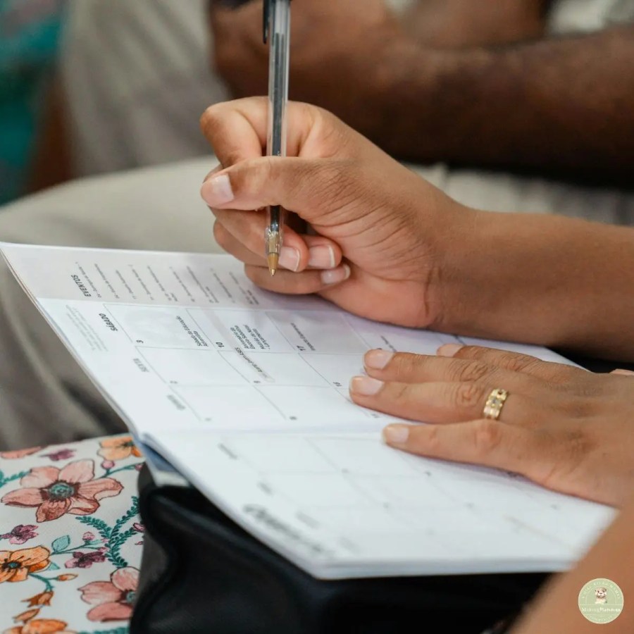 A close-up shot of a person's hands as they write in a planner or calendar with a black ballpoint pen. The person has a gold ring on their right ring finger. The planner is open to a monthly view with some text visible. The background is blurred, showing a floral pattern on the left and a glimpse of another person.