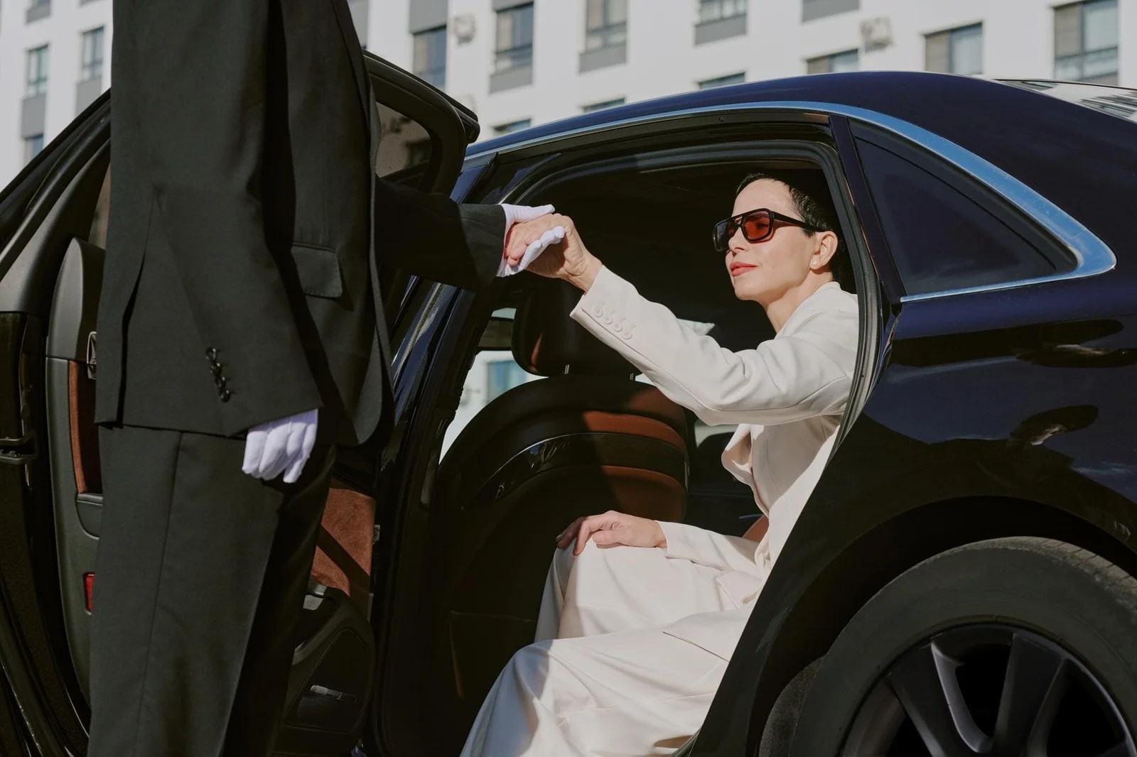 Woman in white dress and sunglasses getting into a luxury black car with chauffeur.