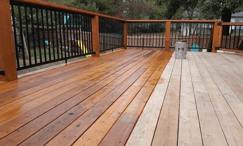 A wooden deck showing a contrast between freshly stained and untreated wood sections, with a railing and some outdoor furniture visible in the background.