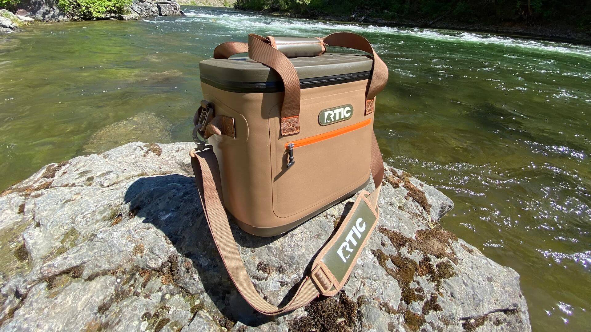 RTIC Ultra-Tough Soft Cooler on a rock near a river with the lid closed.