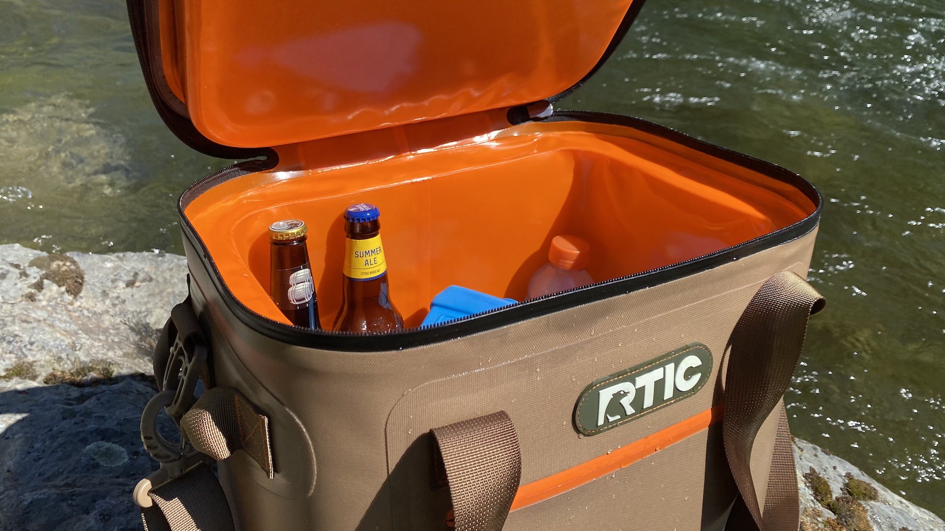 RTIC Ultra-Tough Soft Cooler with lid open and drinks inside on a rock next to a river.