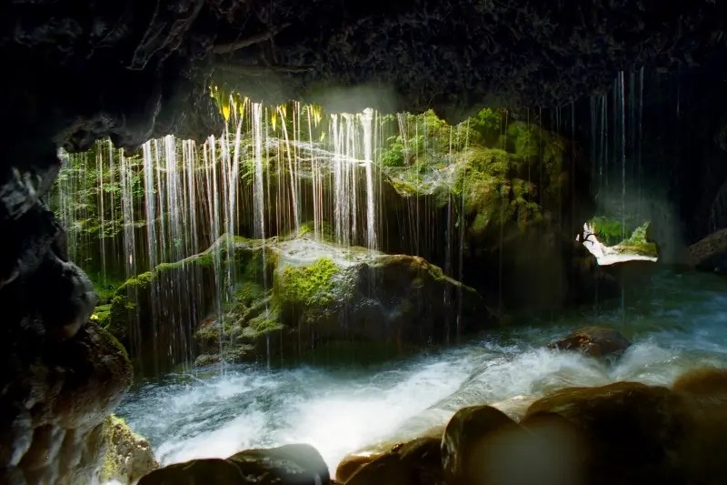Interior del Forat del Bulí con cortina de agua cayendo sobre rocas cubiertas de musgo y corriente de agua clara