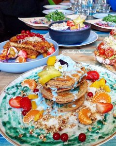 A stack of colourful pancakes topped with cream, mint leaves, and various fruits such as strawberries, oranges, and mango on a decorative plate. In the background, there are bowls of fruits and other dishes, creating a vibrant brunch setting.
