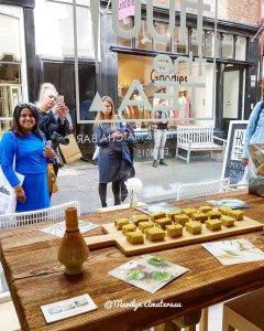 A wooden table displaying a selection of sweets, viewed from inside a café, with two women looking through the window from outside.