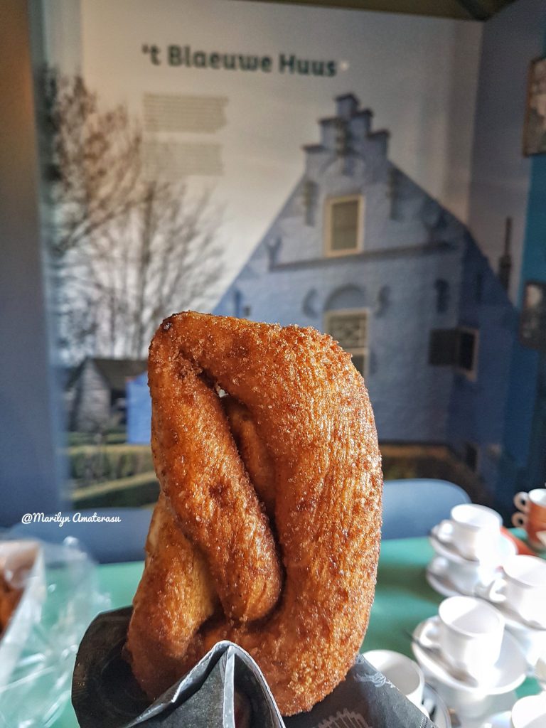 A close-up of a twisted pastry, golden brown and crispy, placed on a black wrapper. In the background, a blue building is partially visible, accompanied by informational text.