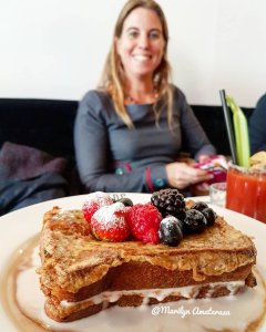 A plate of French toast topped with fresh berries, including raspberries, blackberries, and blueberries, with a woman smiling in the background.