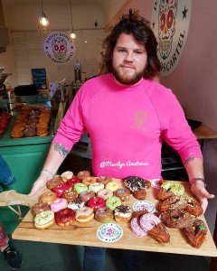 A man in a bright pink shirt holds a wooden board filled with an assortment of colourful doughnuts and pastries, standing in a café with additional doughnuts displayed in the background.