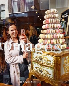 A woman holding a phone takes a selfie in front of a display featuring a tall stack of macarons atop an ornate cabinet, with reflections of passersby in the window.