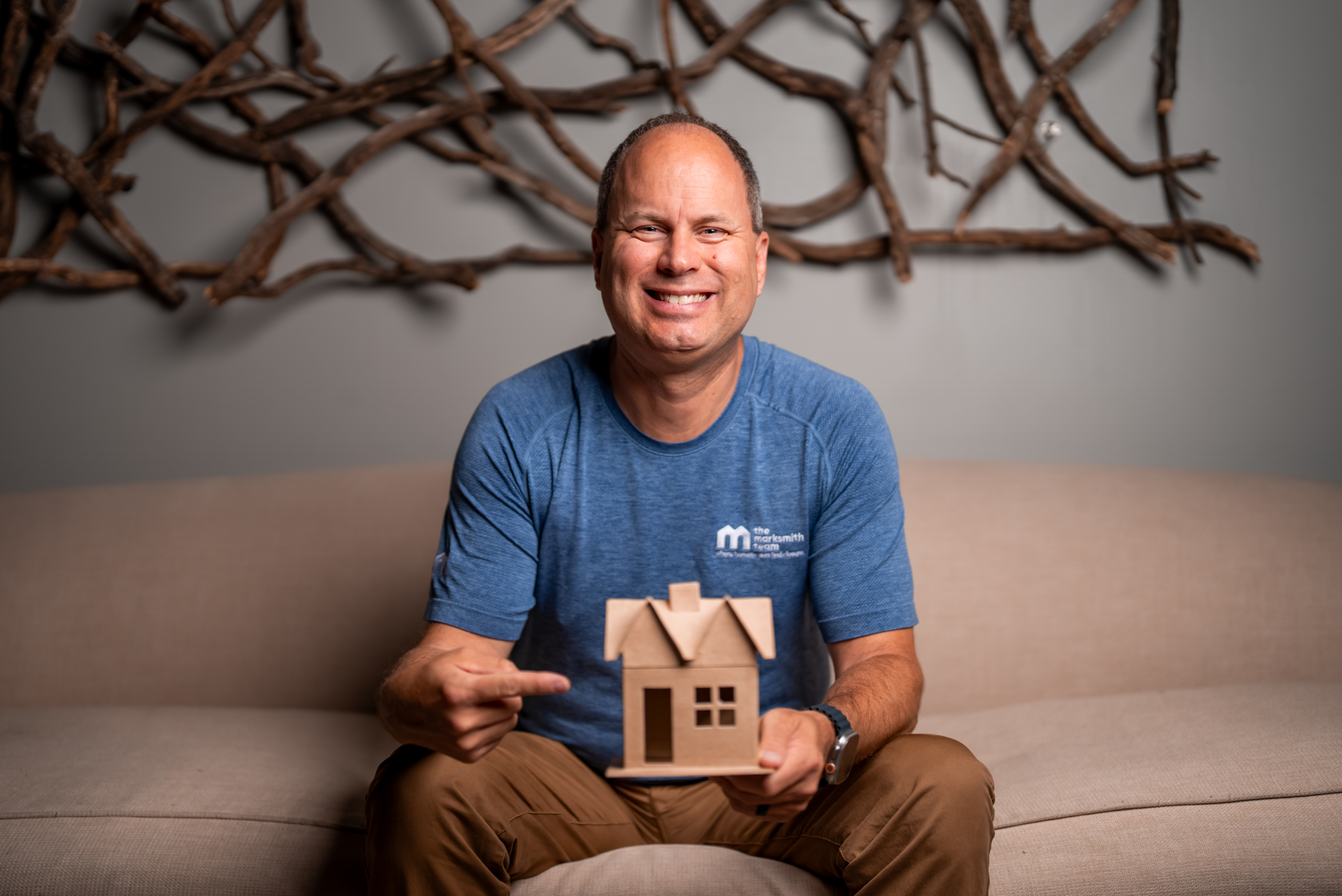 Mark Smith smiling, seated in a The Mark Smith Team shirt holding a house.  