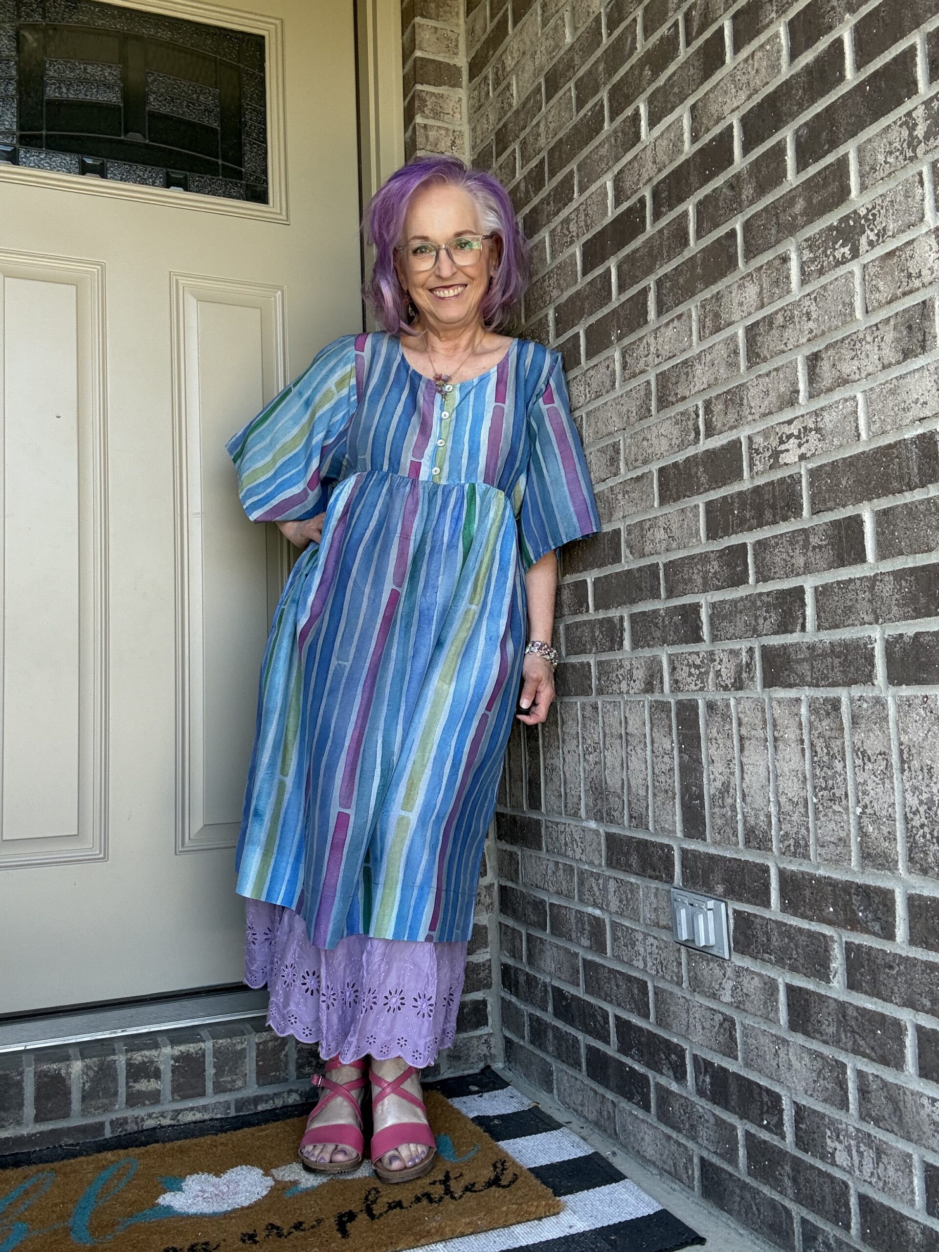 Smiling woman with purple hair and glasses standing at a front door, wearing a blue striped dress and pink sandals beside a brick wall.