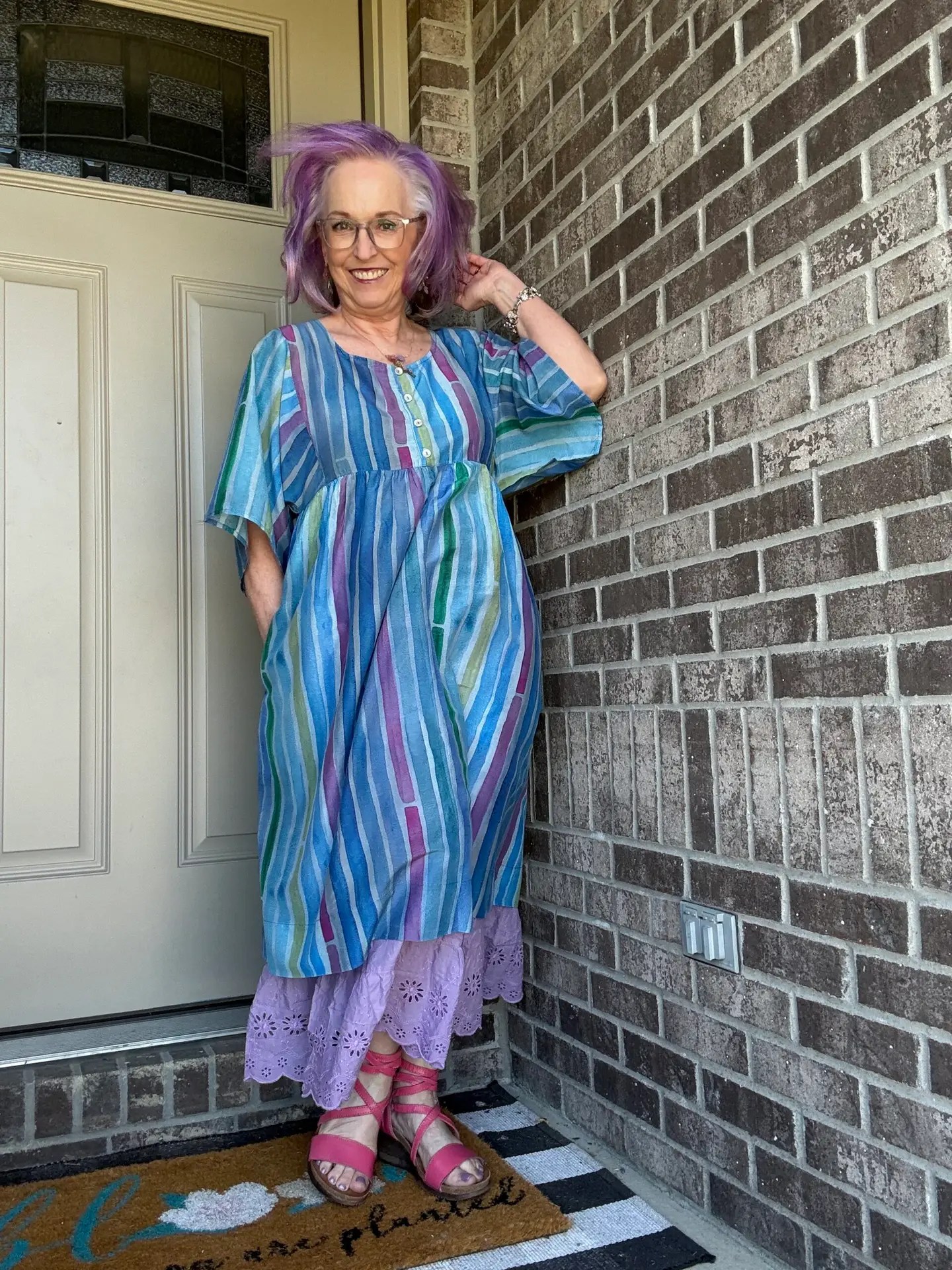 Woman with purple hair wearing a blue striped dress, standing at a door on a brick porch, smiling at the camera.