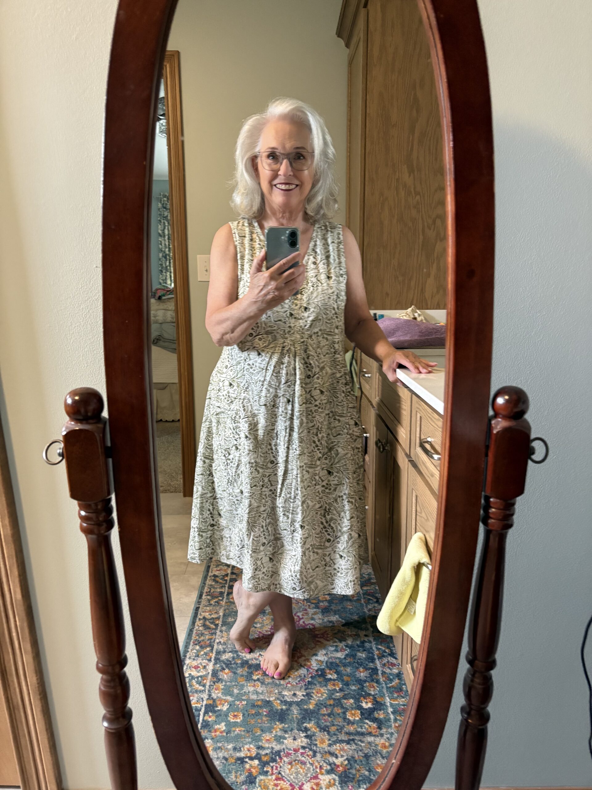 Smiling elderly woman in a light patterned sleeveless dress takes a selfie in a bedroom mirror with a smartphone.