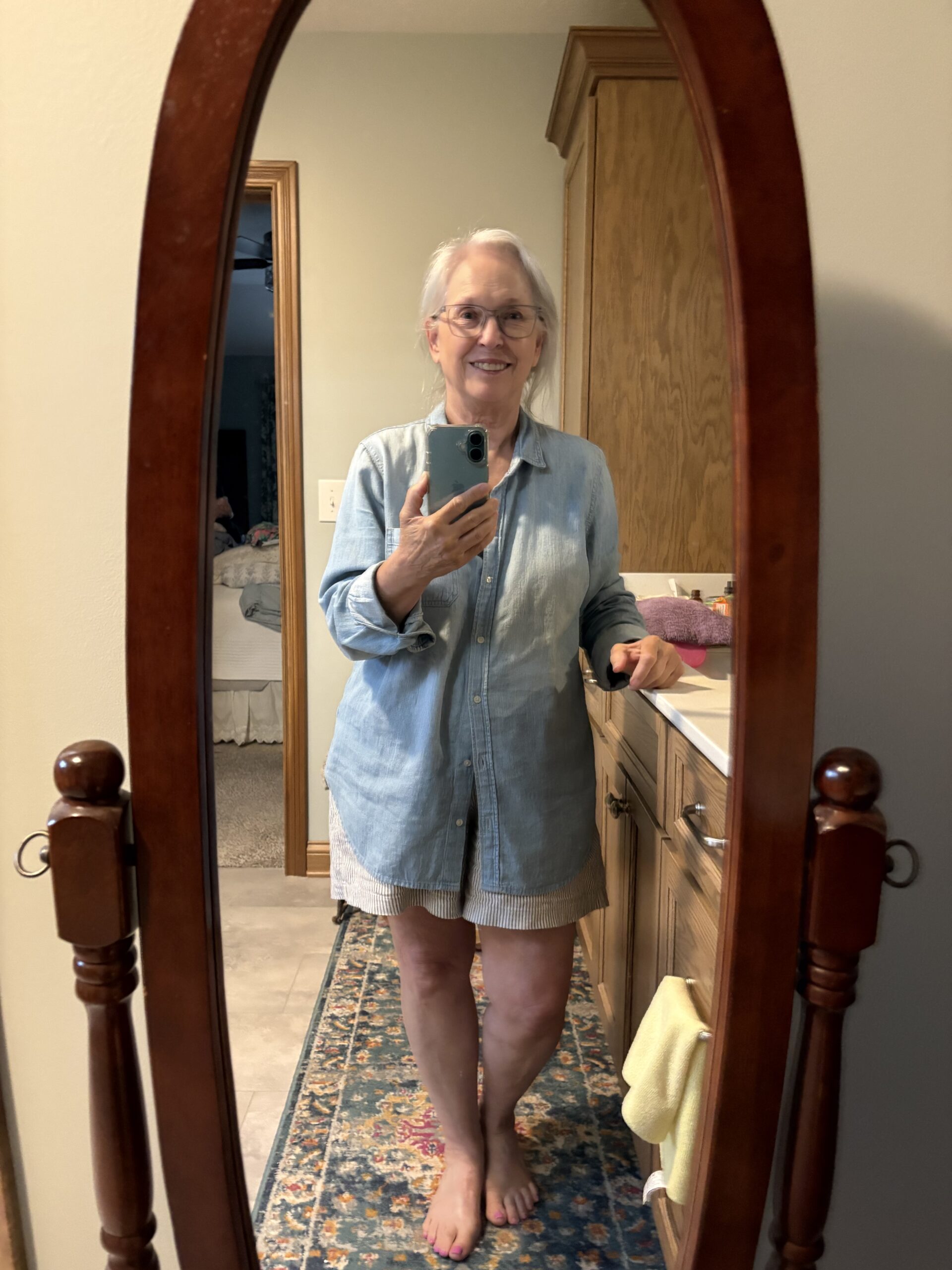 Older woman with gray hair and glasses takes a mirror selfie in a wooden oval frame, wearing a light denim shirt and striped shorts, barefoot.
