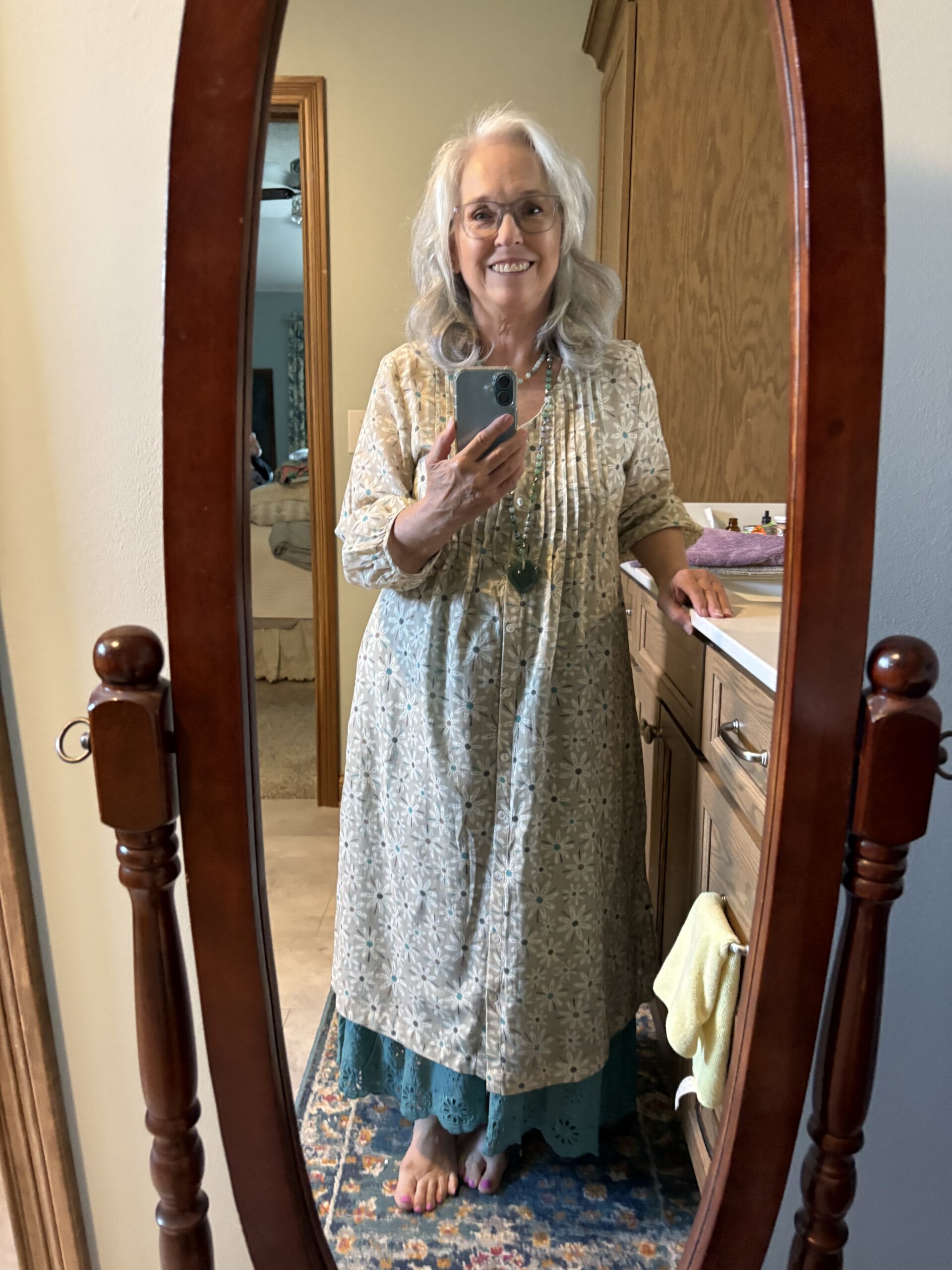 Smiling elderly woman with gray hair and glasses taking a mirror selfie in a bedroom, wearing a light floral dress and turquoise hem, barefoot.