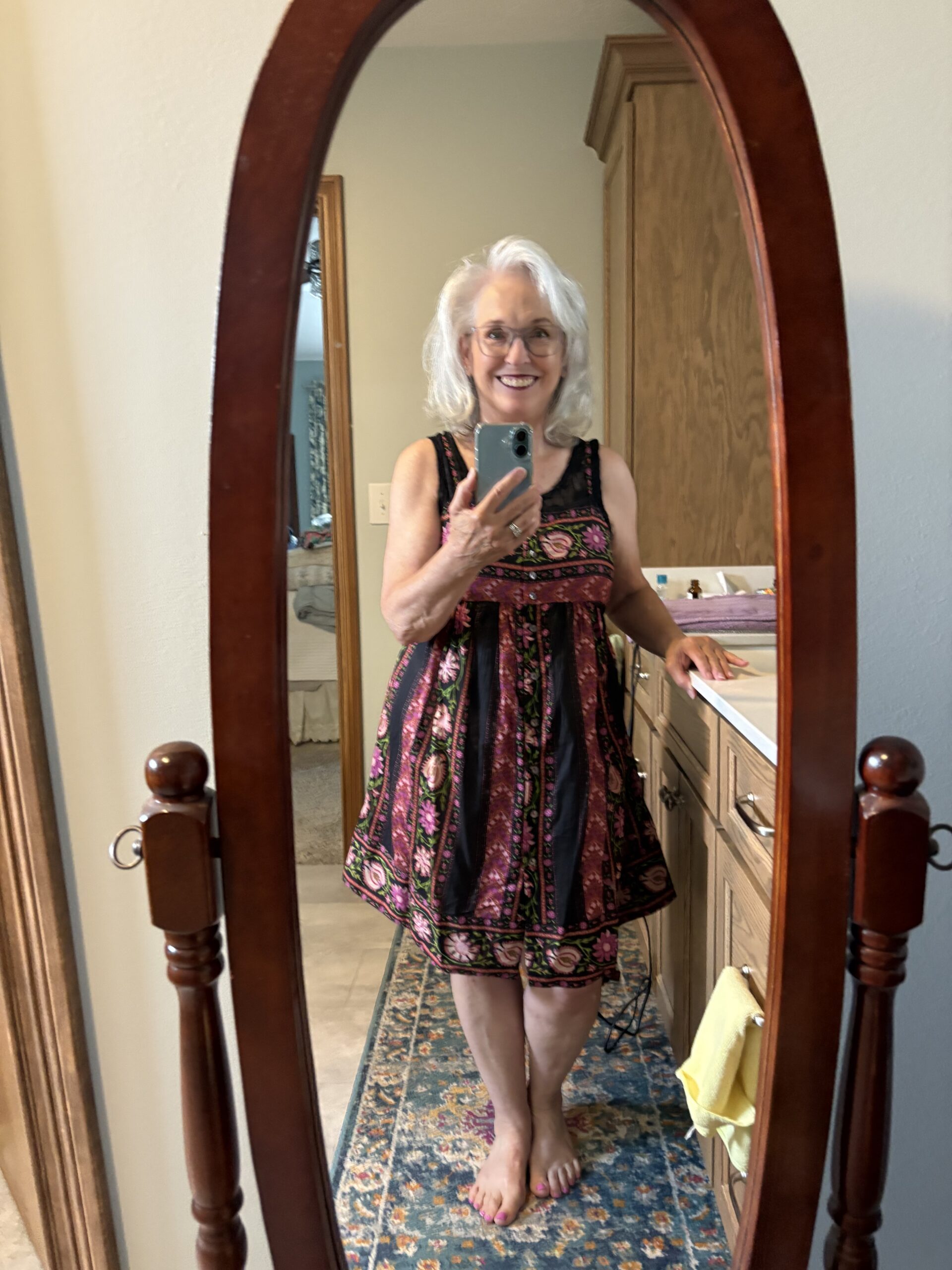 Smiling older woman takes a mirror selfie with a smartphone, wearing a colorful patterned dress in a wooden dresser mirror setting.