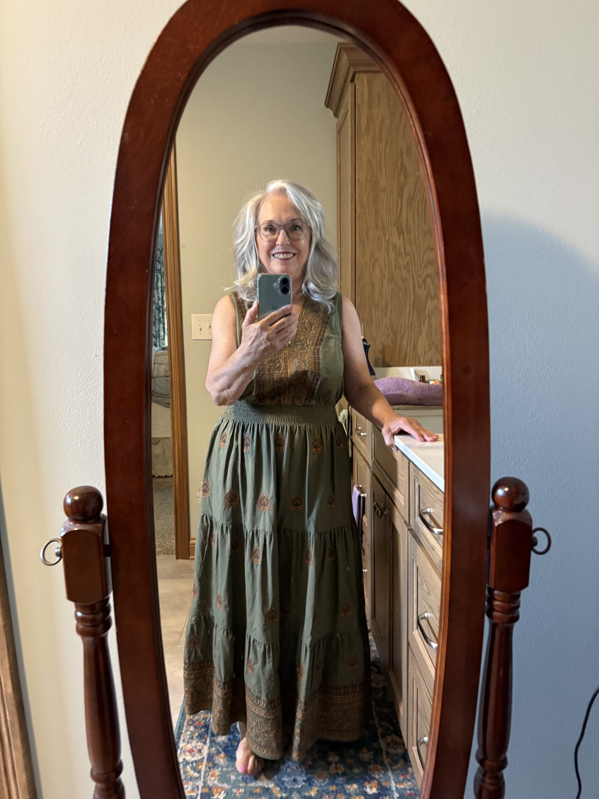 Older woman with gray hair and glasses takes a mirror selfie in a bedroom, wearing a green patterned maxi dress.