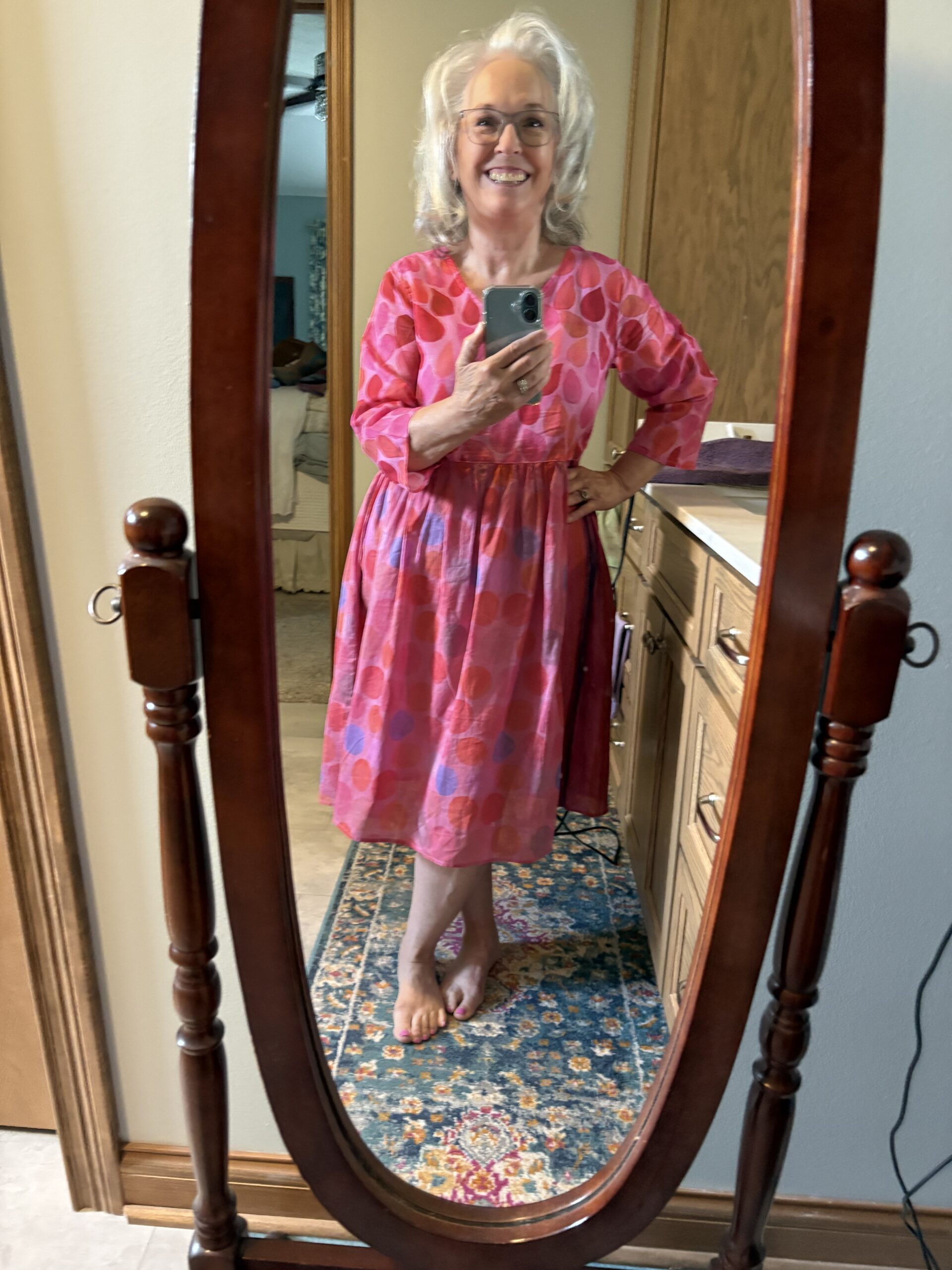 Smiling older woman in a pink patterned dress takes a mirror selfie in a wooden-framed mirror at home, barefoot.