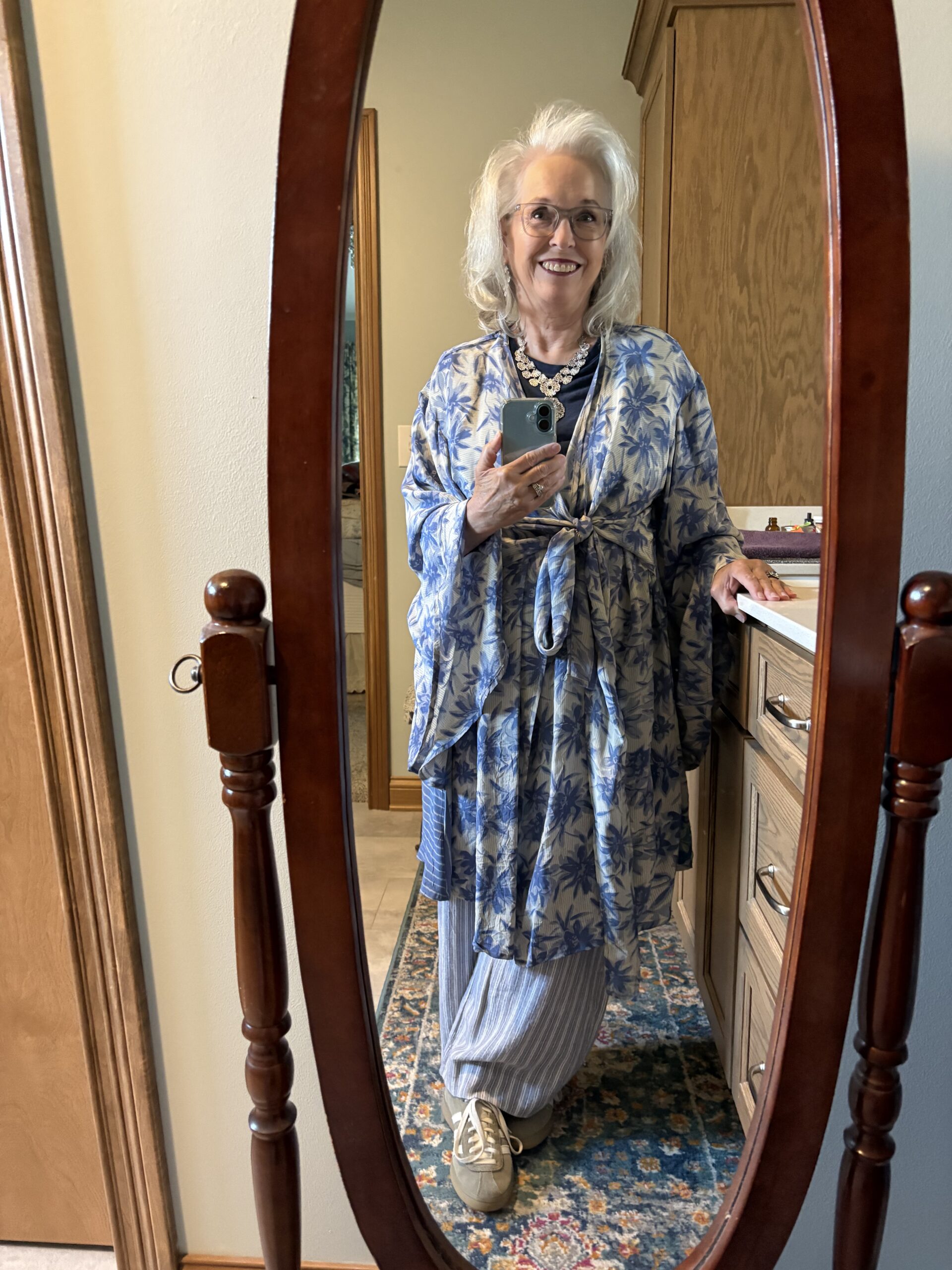 Woman in a blue floral robe posing in a bedroom mirror, holding a smartphone and smiling, with a necklace and glasses visible.