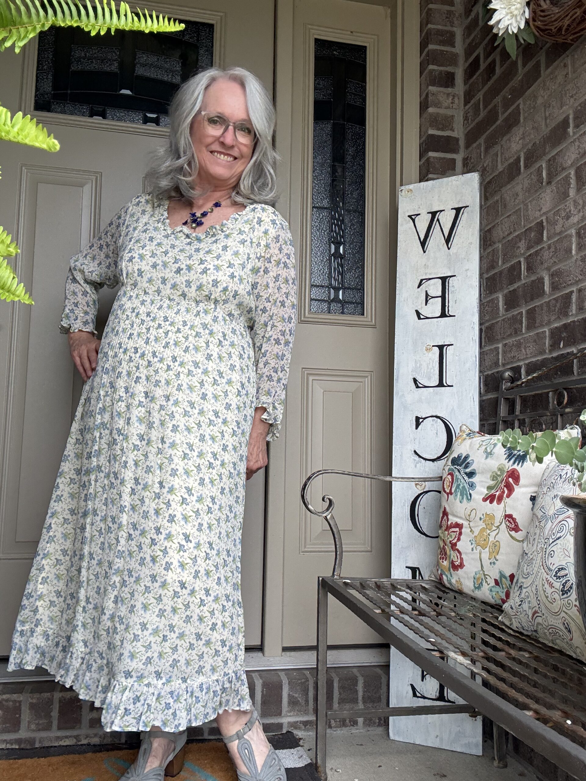 Smiling older woman with gray hair and glasses stands on a front porch in a light blue floral maxi dress, beside a vertical welcome sign reading WELCOME.
