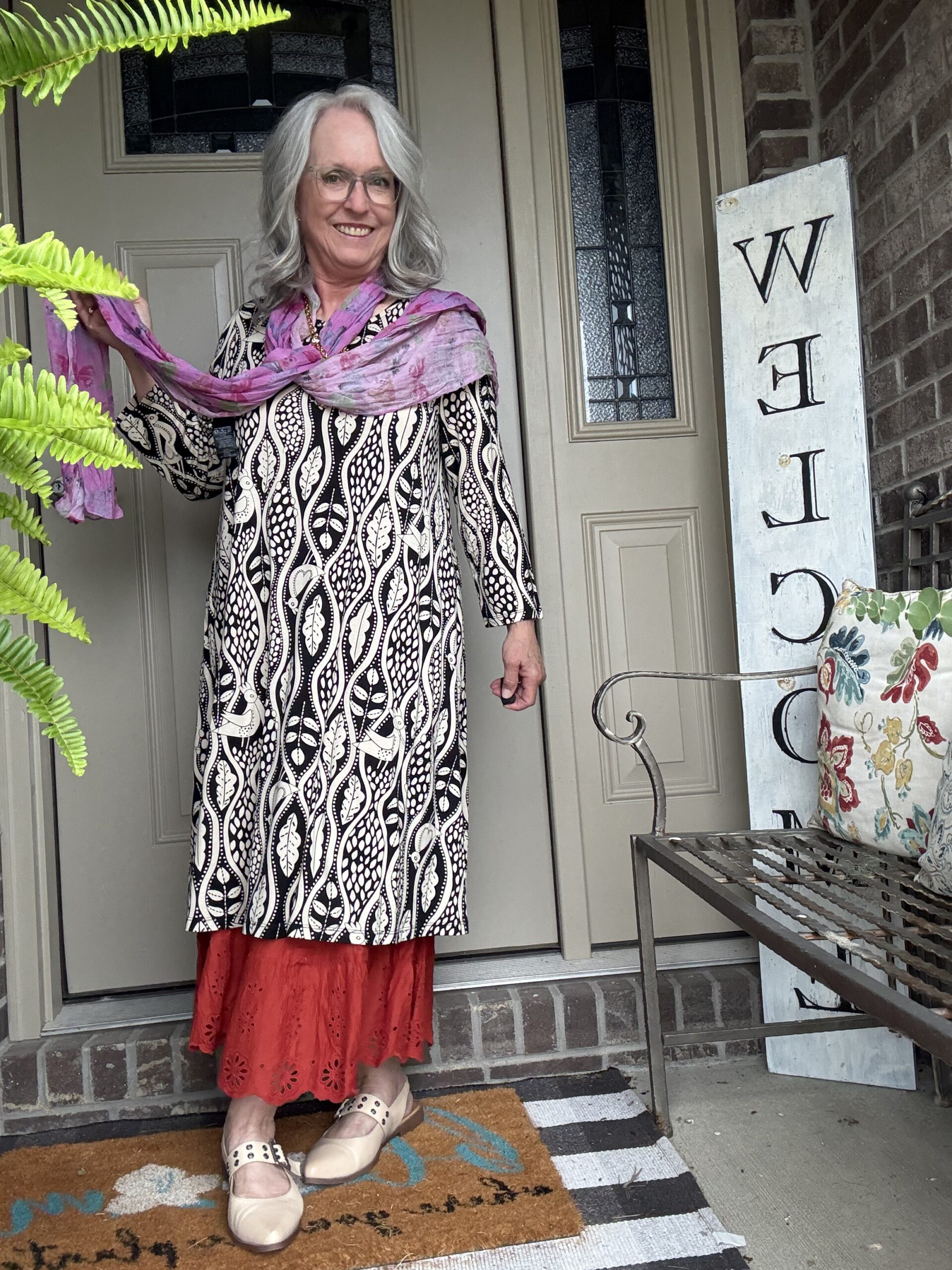 Smiling older woman with gray hair and glasses on a front porch, wearing a black‑and‑white patterned dress with a pink scarf and a red lace hem, beige sandals.
