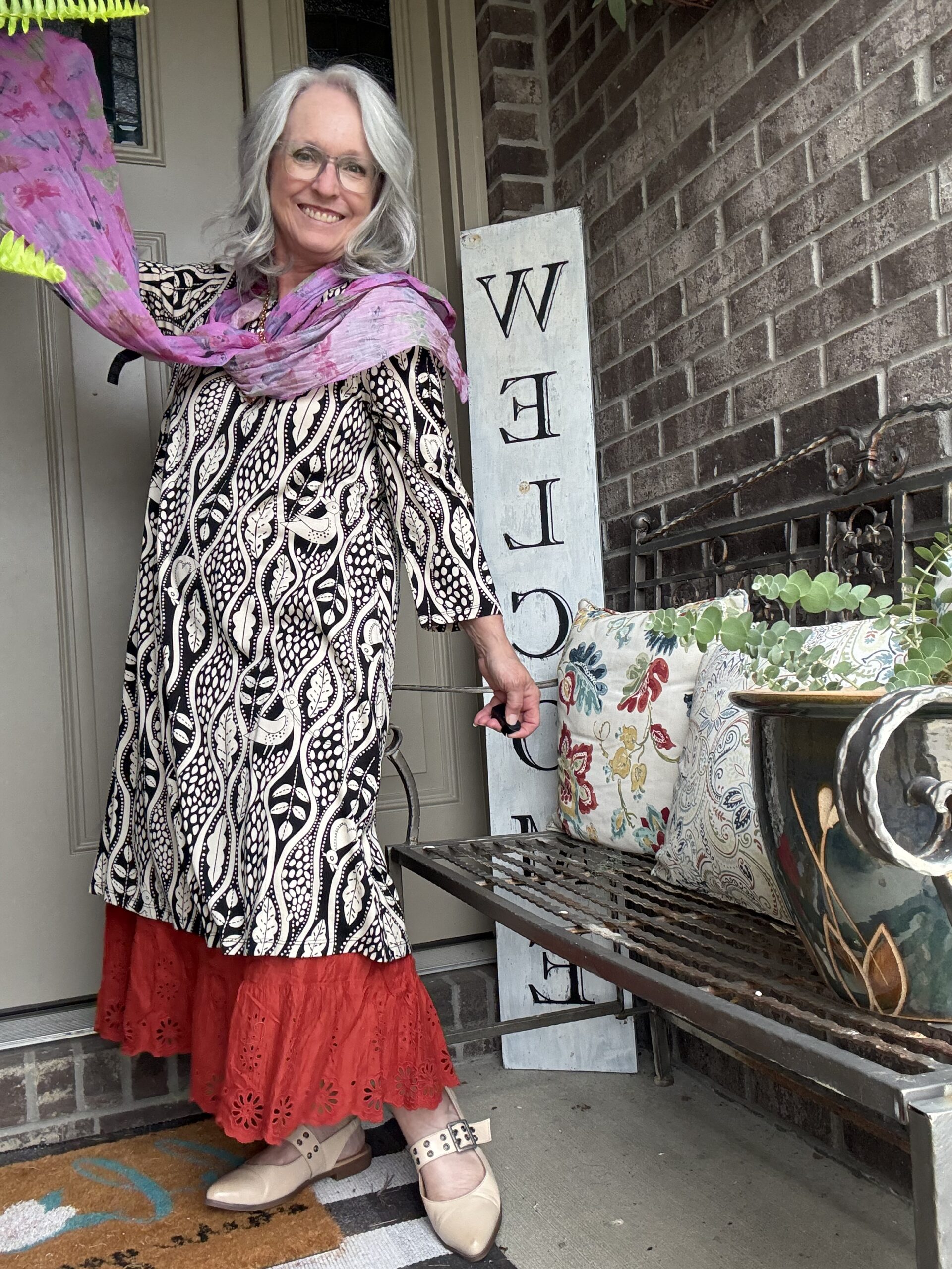 Smiling woman with gray hair and glasses stands at a front door, wearing a black-and-white patterned dress, a pink scarf, and beige shoes, beside a vertical welcome sign.