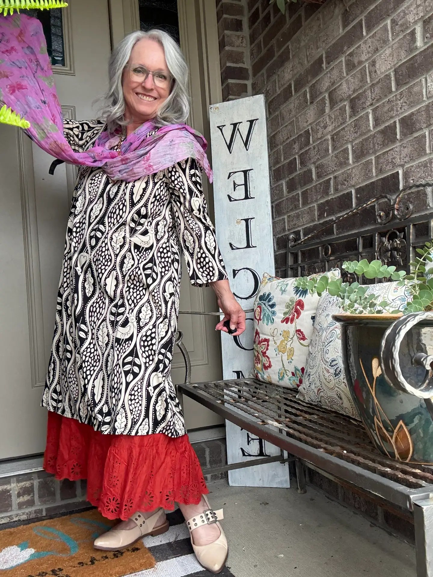 Smiling woman with gray hair and glasses stands at a front door, wearing a black-and-white patterned dress, a pink scarf, and beige shoes, beside a vertical welcome sign.