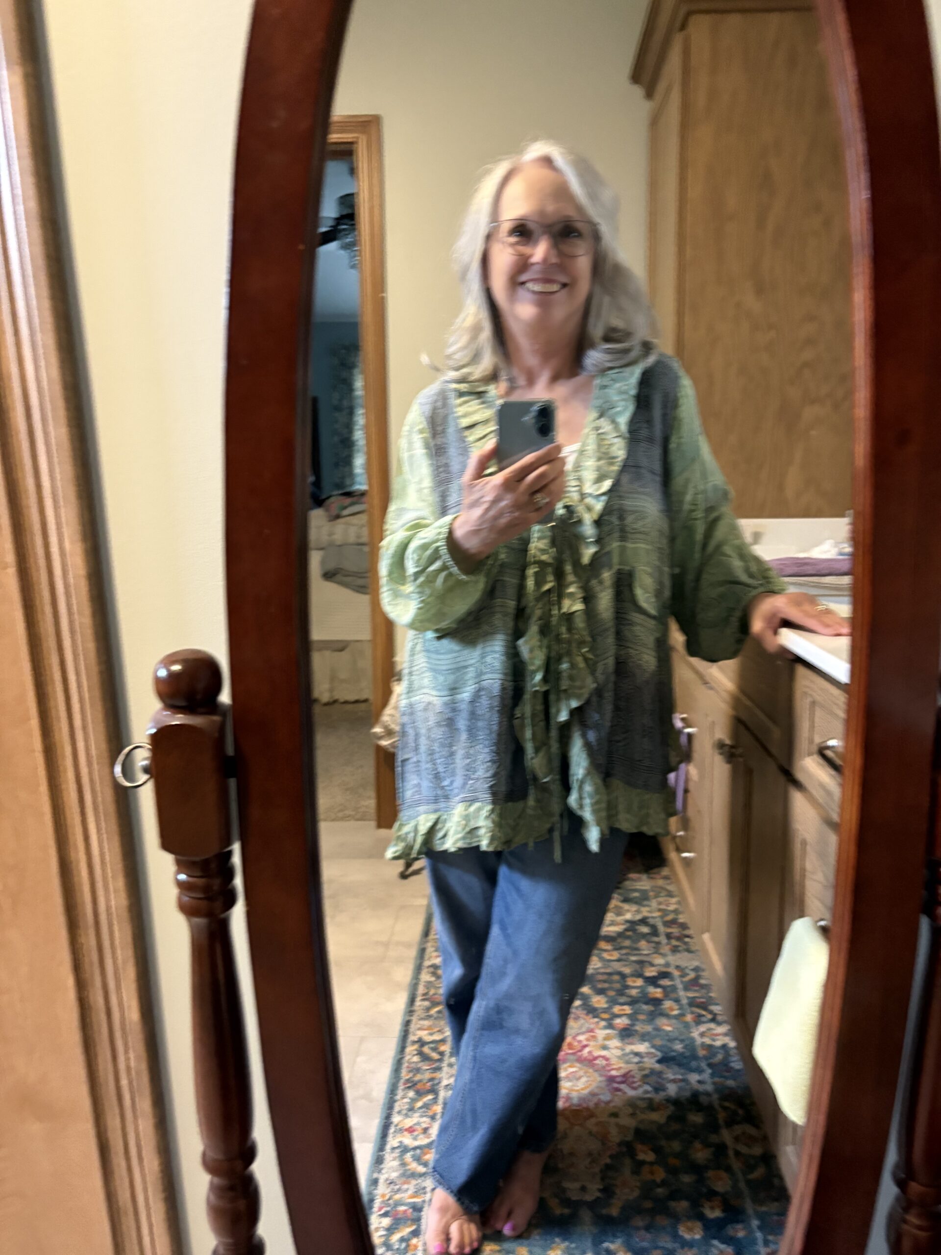 Smiling woman with gray hair taking a mirror selfie, wearing a green patterned blouse and blue jeans.