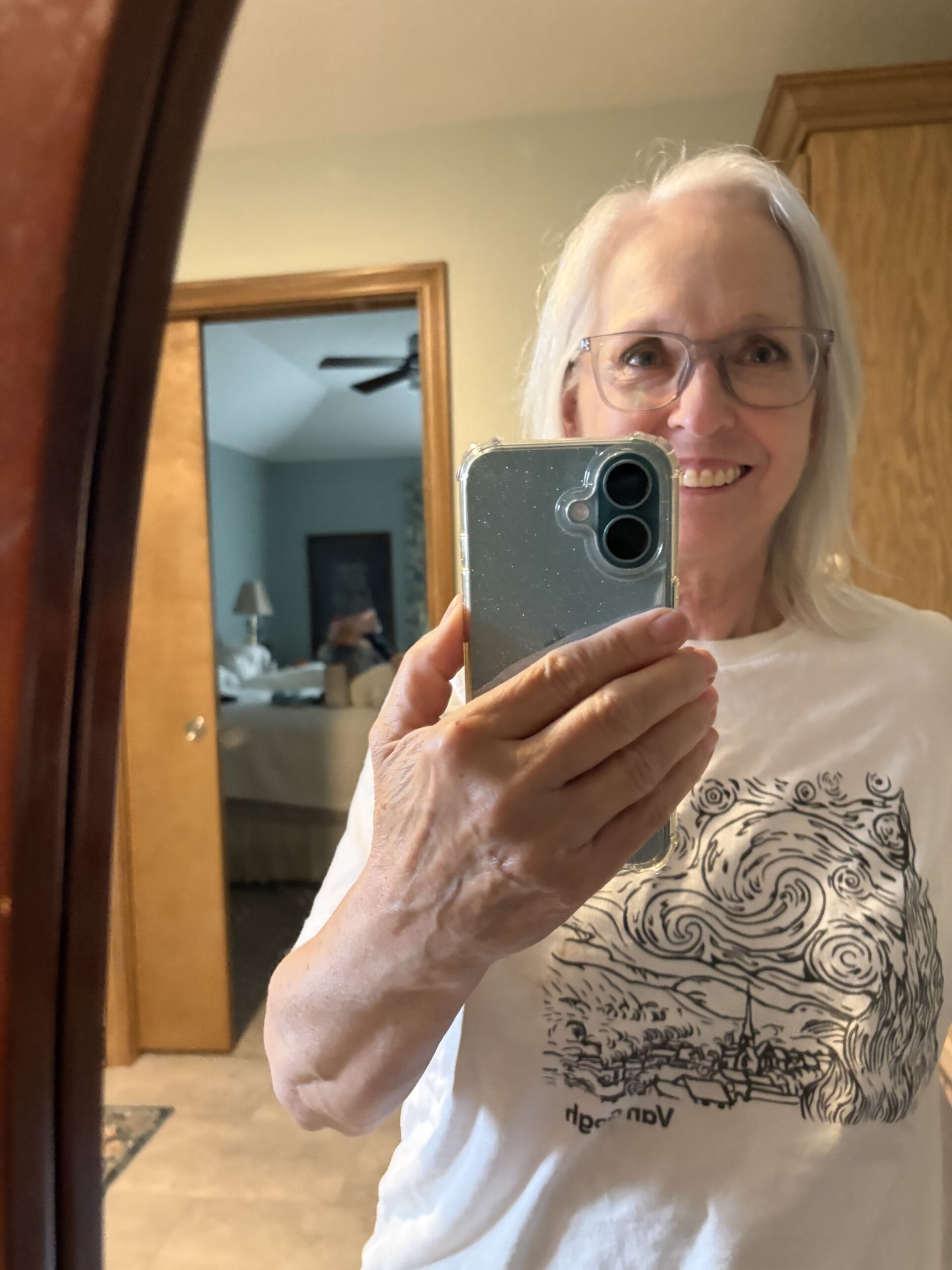 Smiling woman with white hair and glasses taking a mirror selfie with a smartphone in hand.