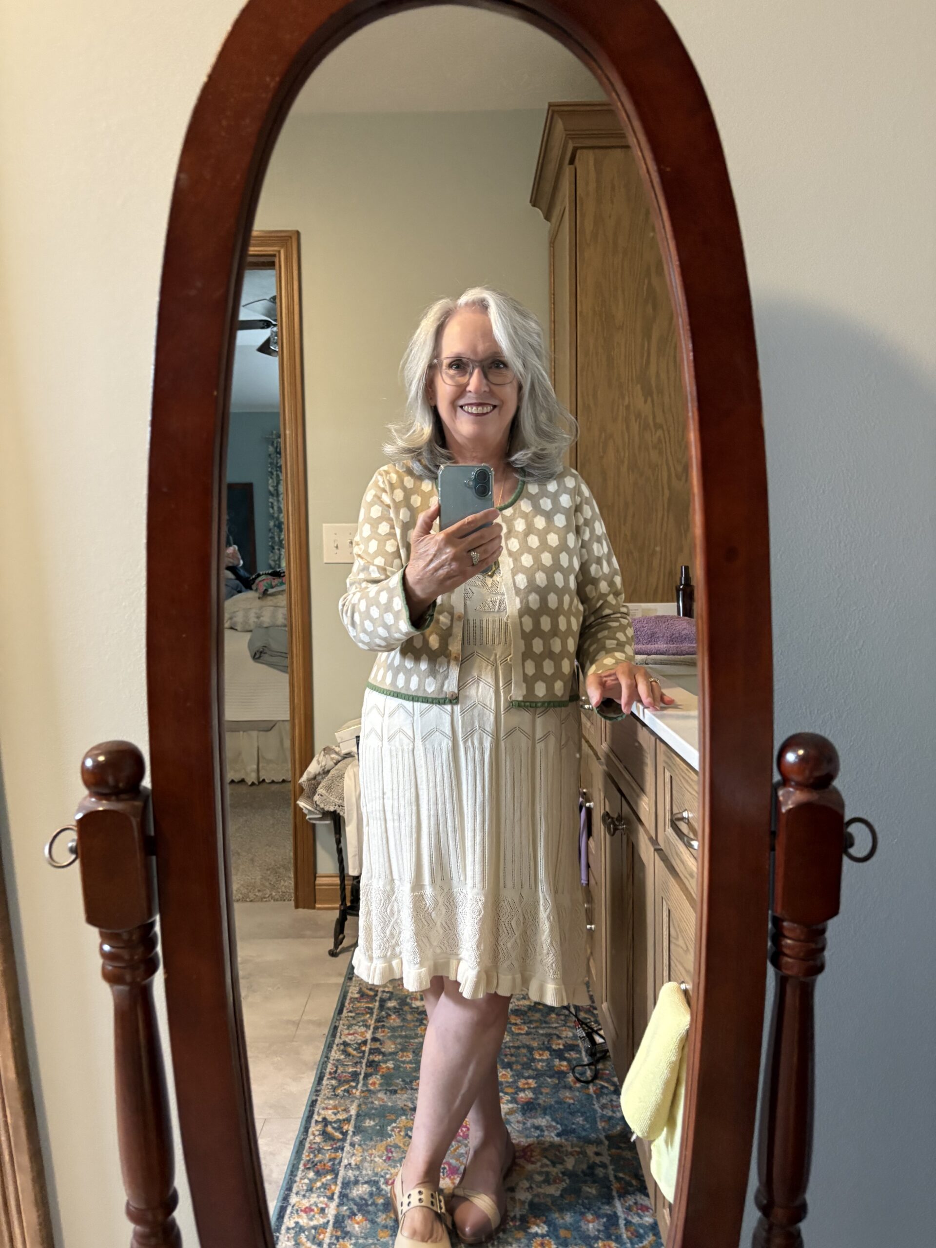 Smiling woman with gray hair taking a mirror selfie in a bedroom, wearing a cream patterned cardigan and dress, holding a smartphone.