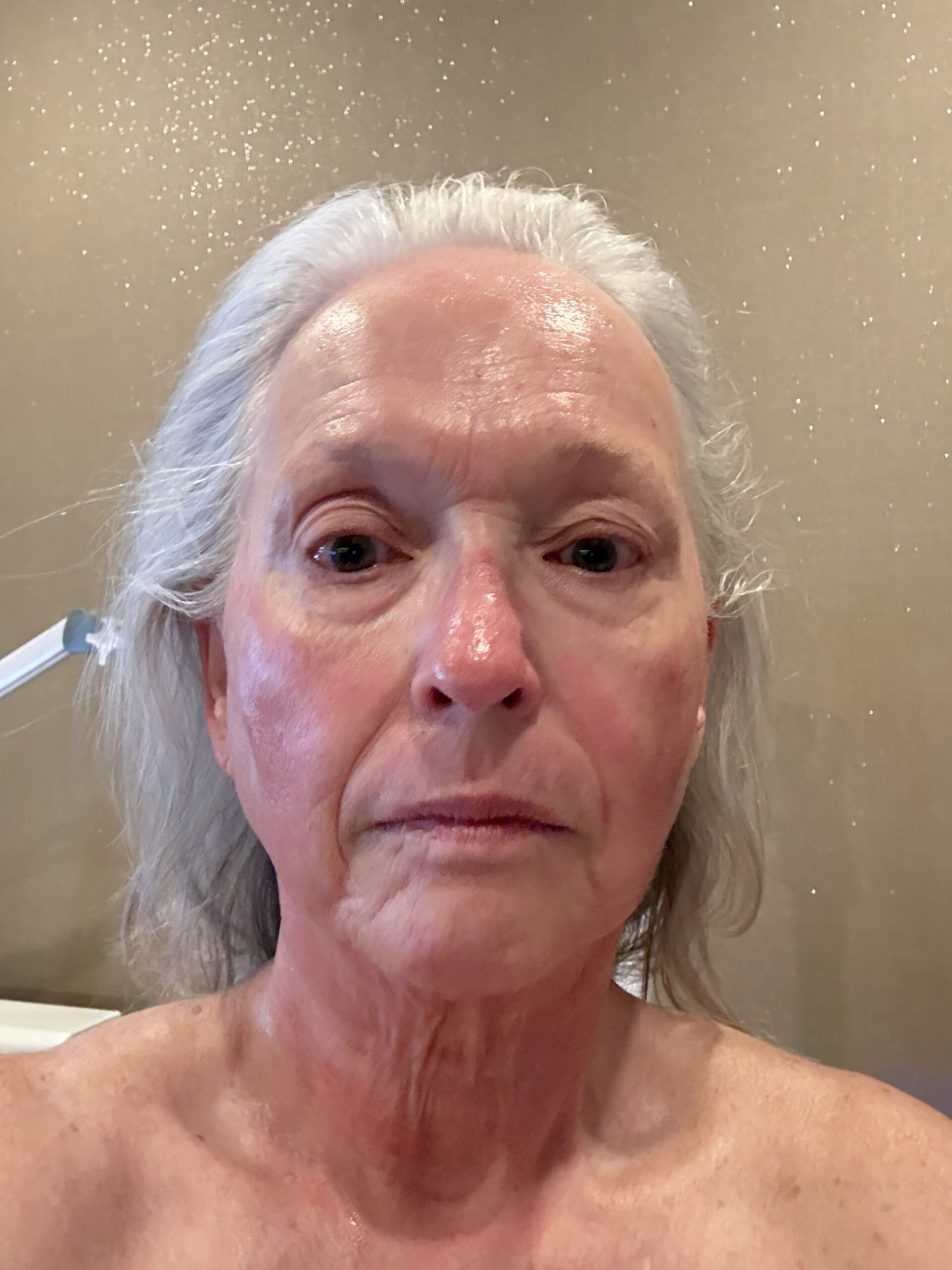 Close-up of an elderly woman's wet face and silver hair, looking at the camera in a bathroom setting.