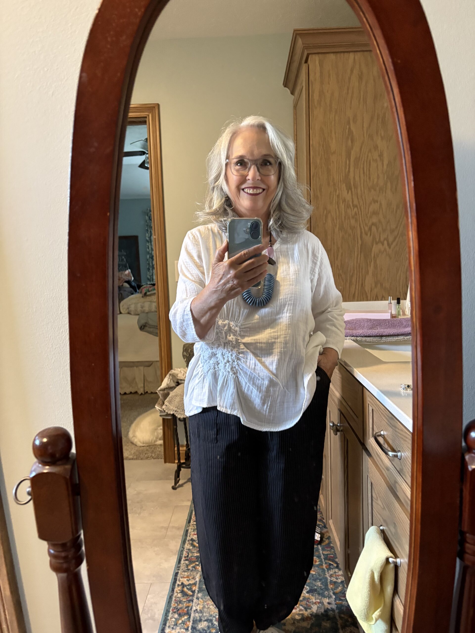Smiling older woman with gray hair and glasses taking a mirror selfie in a wooden arched dresser mirror, wearing a white blouse and dark pinstripe pants with a chunky necklace.