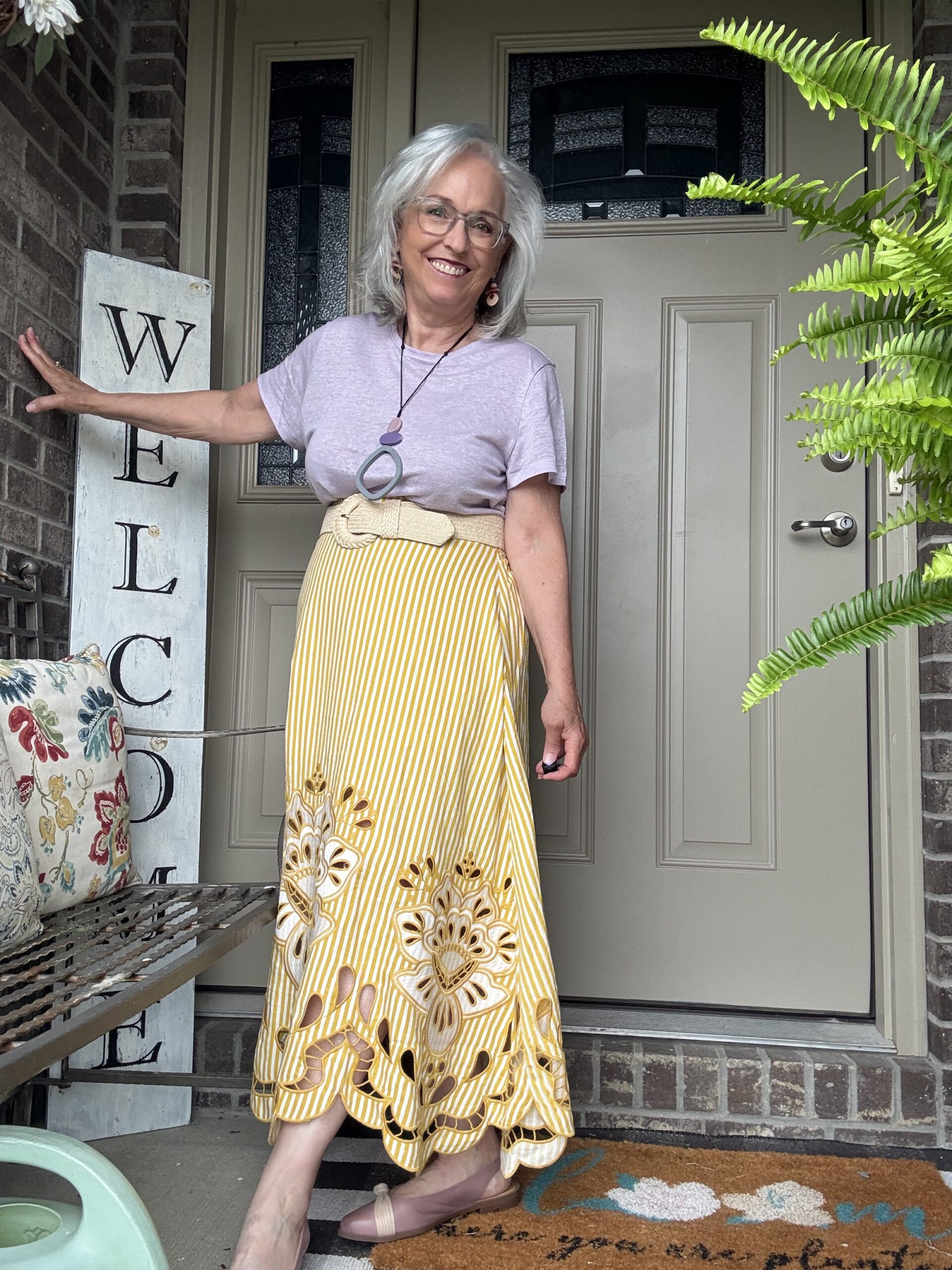 Cheerful older woman in a lavender top and yellow striped skirt stands on a front porch beside a vertical 'WELCOME' sign, smiling at the camera.